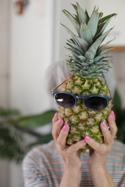 портрет зрелой седой женщины, держащей спелый ананас в солнечных очках перед лицом дома