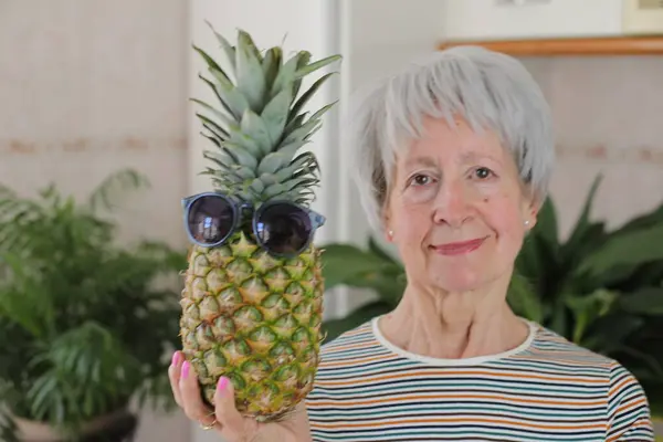 stock image portrait of mature grey haired woman holding pineapple with sunglasses at home