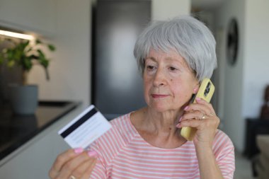 Skeptical looking senior woman holding credit card during phone call clipart