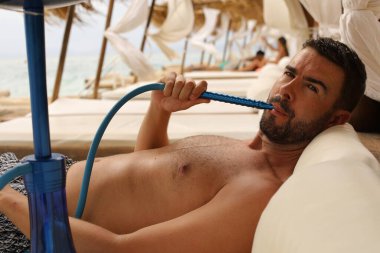 Yakışıklı adam plajda nargileyle yatıyor.