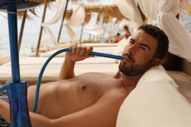 Yakışıklı adam plajda nargileyle yatıyor.