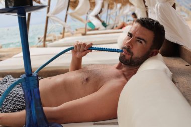 Yakışıklı adam plajda nargileyle yatıyor.