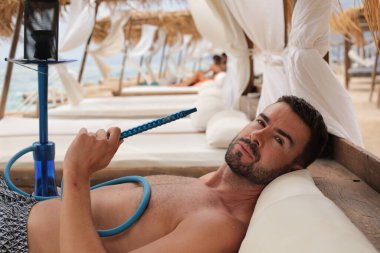 Yakışıklı adam plajda nargileyle yatıyor.