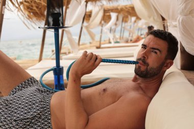 Yakışıklı adam plajda nargileyle yatıyor.