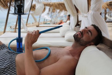 Yakışıklı adam plajda nargileyle yatıyor.