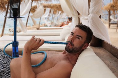 Yakışıklı adam plajda nargileyle yatıyor.