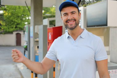 Genç yakışıklı adam gülümsüyor, istasyonda araba pompası tutuyor.