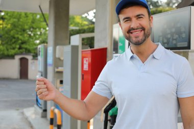 Genç yakışıklı adam gülümsüyor, istasyonda araba pompası tutuyor.