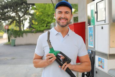 Genç yakışıklı adam gülümsüyor, istasyonda araba pompası tutuyor.