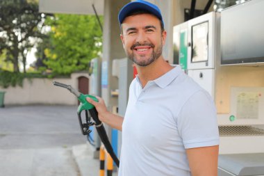 Genç yakışıklı adam gülümsüyor, istasyonda araba pompası tutuyor.