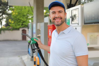 Genç yakışıklı adam gülümsüyor, istasyonda araba pompası tutuyor.