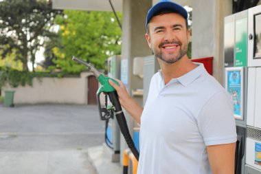 Genç yakışıklı adam gülümsüyor, istasyonda araba pompası tutuyor.