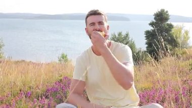 Mutlu genç adam göl kenarında elma yiyor.