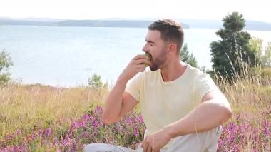 Mutlu genç adam göl kenarında elma yiyor.