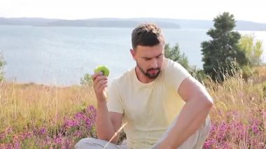 Mutlu genç adam göl kenarında elma yiyor.