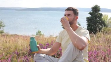Termoslu genç bir adam Çiçekli çayır ve göllü bir çay