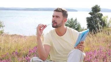 Sarı gömlekli bir adam su kenarında defter yazıyor.