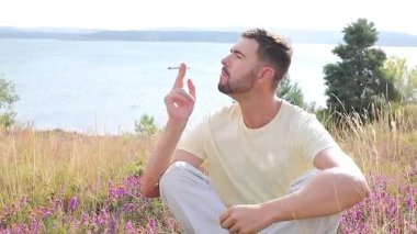 Genç adam, gölün kenarında elinde çiçeklerle sigara içiyor.