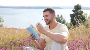 Sarı gömlekli bir adam su kenarında defter yazıyor.