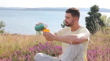 Termoslu genç bir adam Çiçekli çayır ve göllü bir çay