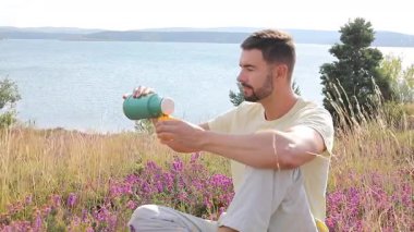 Termoslu genç bir adam Çiçekli çayır ve göllü bir çay