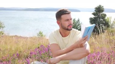 Sarı tişörtlü bir adam arka planda manzaralı açık havada defter okuyor..