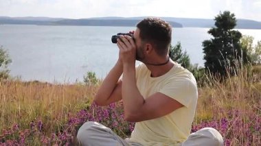 Adam dışarıda oturuyor, elinde bir çift siyah dürbün tutuyor.