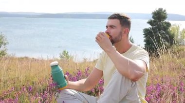 Termoslu genç bir adam Çiçekli çayır ve göllü bir çay