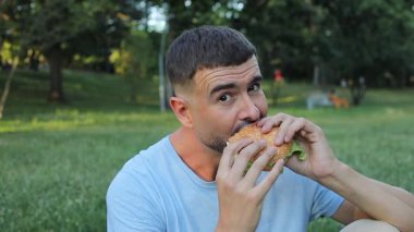 Genç yakışıklı adam parkta hamburger yiyor..