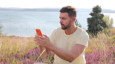 Genç adam göl kenarında akıllı telefon kullanıyor.