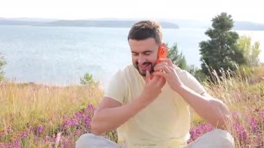 Genç, yakışıklı, sakallı, sıradan kıyafetler giymiş, elinde cep telefonuyla çimlerin üzerinde oturan bir adam..