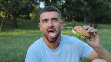 Genç yakışıklı adam parkta hamburger yiyor..