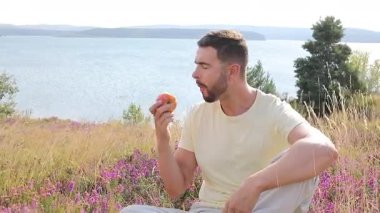 Genç Adam Göl kenarındaki Çimenli bir alanda Red Apple 'dan bir ısırık alıyor..