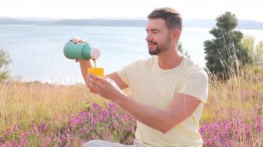 Termoslu genç bir adam Çiçekli çayır ve göllü bir çay