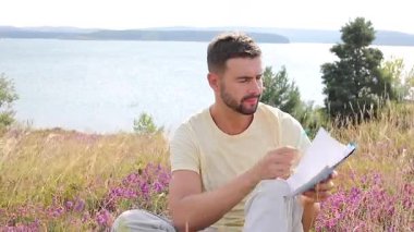 Sarı tişörtlü adam dışarıda, arka planı manzaralı deftere yazıyor..