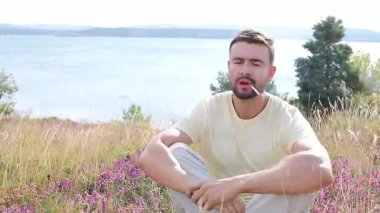 Genç adam, gölün kenarında elinde çiçeklerle sigara içiyor.
