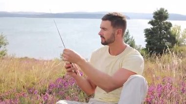 Bir adam bir göl kenarındaki yamaçta oturuyor, elinde radyo var.