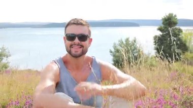 Güneş gözlüklü mutlu genç adam Grassy Hill 'de oturuyor. 