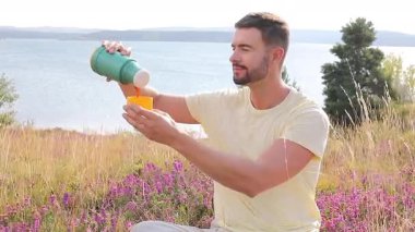 Termoslu genç bir adam Çiçekli çayır ve göllü bir çay