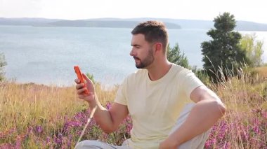 Tepedeki çimlerde otururken akıllı telefon kullanan mutlu bir adam.