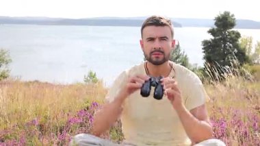 Adam dışarıda oturuyor, elinde bir çift siyah dürbün tutuyor.