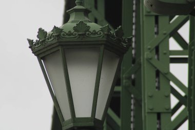 An intricately designed green street lamp stands on a bridge, showcasing decorative elements and ambient lighting, surrounded by industrial structures against a cloudy sky. clipart