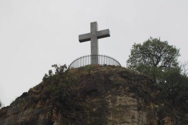 A prominent metallic cross stands atop a rocky outcrop, surrounded by lush vegetation, under a cloudy sky, creating a peaceful atmosphere in nature. clipart