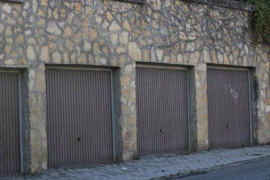 A row of four closed garage doors sits neatly against a rugged stone wall. The sunlight highlights the textured surface, creating a calm, urban atmosphere. clipart