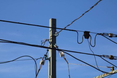 Elektrik direkleri ve elektrik hatları parlak mavi bir gökyüzü boyunca uzanıyor. Bu da temel elektrik altyapısını gösteriyor. Kompozisyon modern enerji dağılımının karmaşıklığını vurgular.