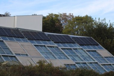 Güneş panelleri, ağaçlar ve mavi gökyüzüyle çevrili çağdaş bir binanın eğimli çatısını kaplayarak sürdürülebilir enerji kaynaklarının önemini vurguluyor..