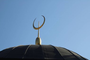 Görüntüde, parlak mavi gökyüzünün altında aydınlanan ve İslam kültürünü sembolize eden bir cami kubbesinin tepesindeki altın hilal ve yıldız yer alıyor..