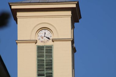 Bir saat kulesi parlak mavi gökyüzüne karşı dimdik durur, karmaşık mimari ayrıntıları ve canlı renkleri ortaya çıkarır. Gösterilen zaman, tarihi bir ortamda gün ortası olduğunu gösteriyor.
