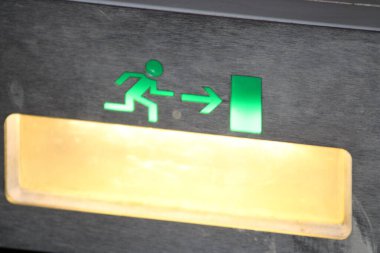 A vibrant green emergency exit sign shows a running figure and an arrow pointing to the right, guiding individuals towards safety. The background is dark, enhancing visibility. clipart