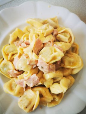 İçinde peynir ve jambon dolu yumuşak makarna bulunan bol kremalı bir tortellini tabağı. Yemek iştah açıcı görünüyor ve basit ve zarif bir şekilde sunuluyor..